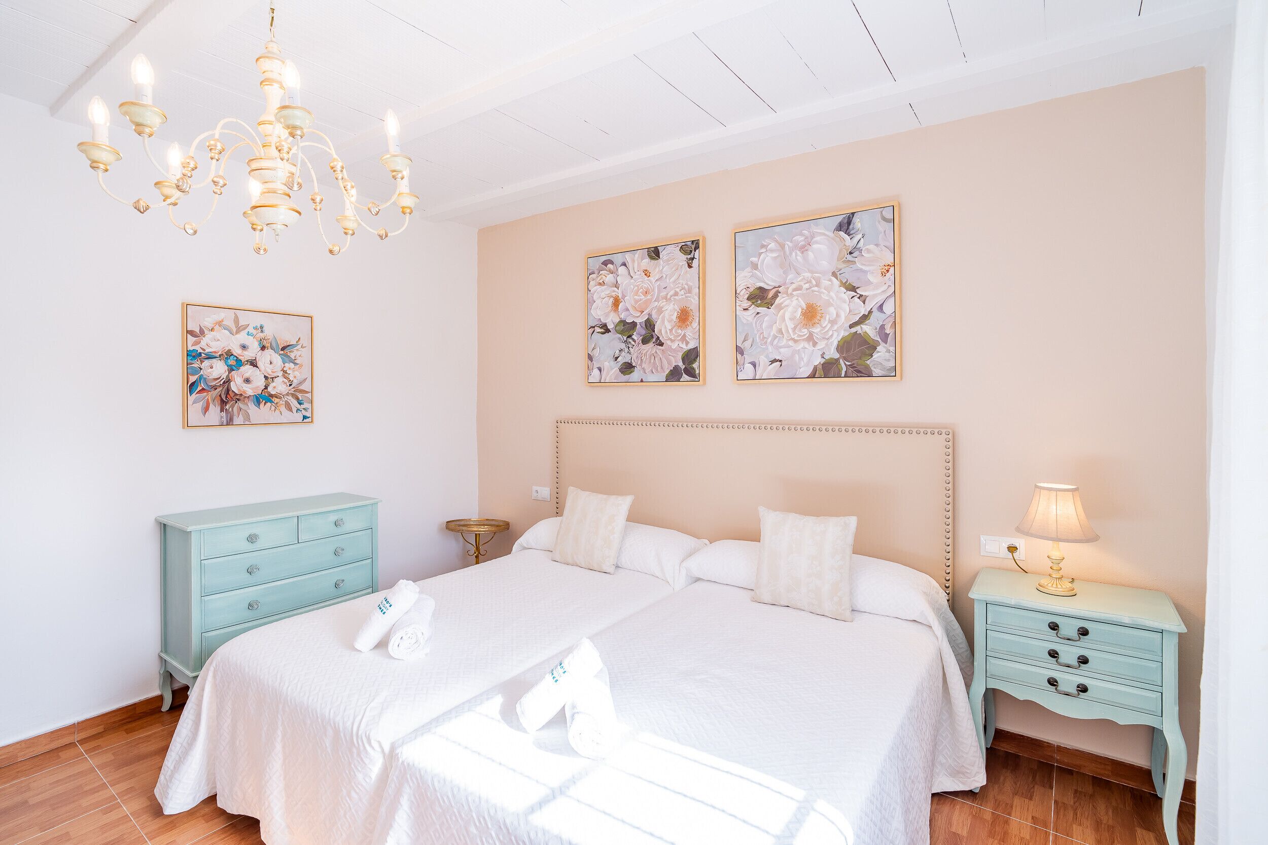 Una cama doble con sábanas blancas, un aparador azul claro, una mesita de noche con lámpara y obras de arte con motivos florales en las paredes.