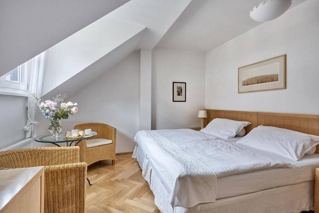 A bedroom with a bed, a chair, a small table with a vase, and a picture on the wall.