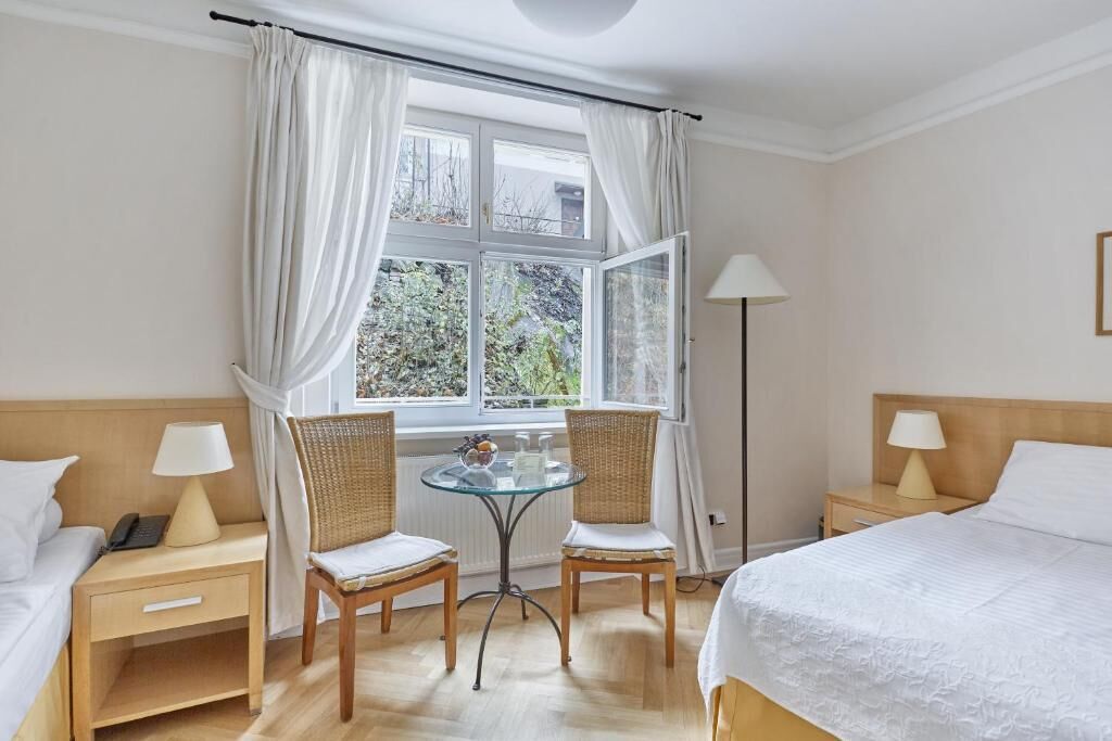 Habitación de hotel con una cama, dos mesitas de noche, una mesa con sillas, una lámpara de pie y un ventanal con cortinas.