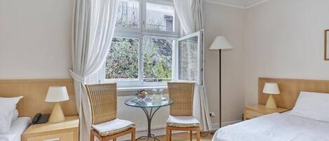 Habitación de hotel con una cama, dos mesitas de noche, una mesa con sillas, una lámpara de pie y un ventanal con cortinas.