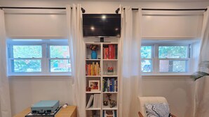 A room with a bookshelf, a TV, a small table with a suitcase, a couch, and two windows with curtains. - 2-bedroom Rowhome w/ AC in South Philly 10 min to Plane/Train & 5 min to Stadium (Philadelphia)