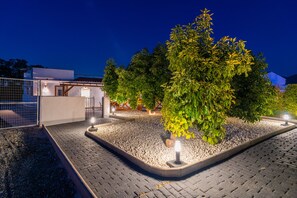 Ein Außenbereich bei Nacht mit einem gepflasterten Weg, einem eingezäunten Bereich und einem Baum, der von einer Lampe beleuchtet wird.