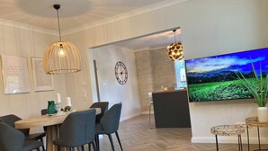 Un comedor moderno con una mesa de madera, sillas oscuras, luces colgantes, un reloj de pared y un televisor grande que muestra un paisaje.