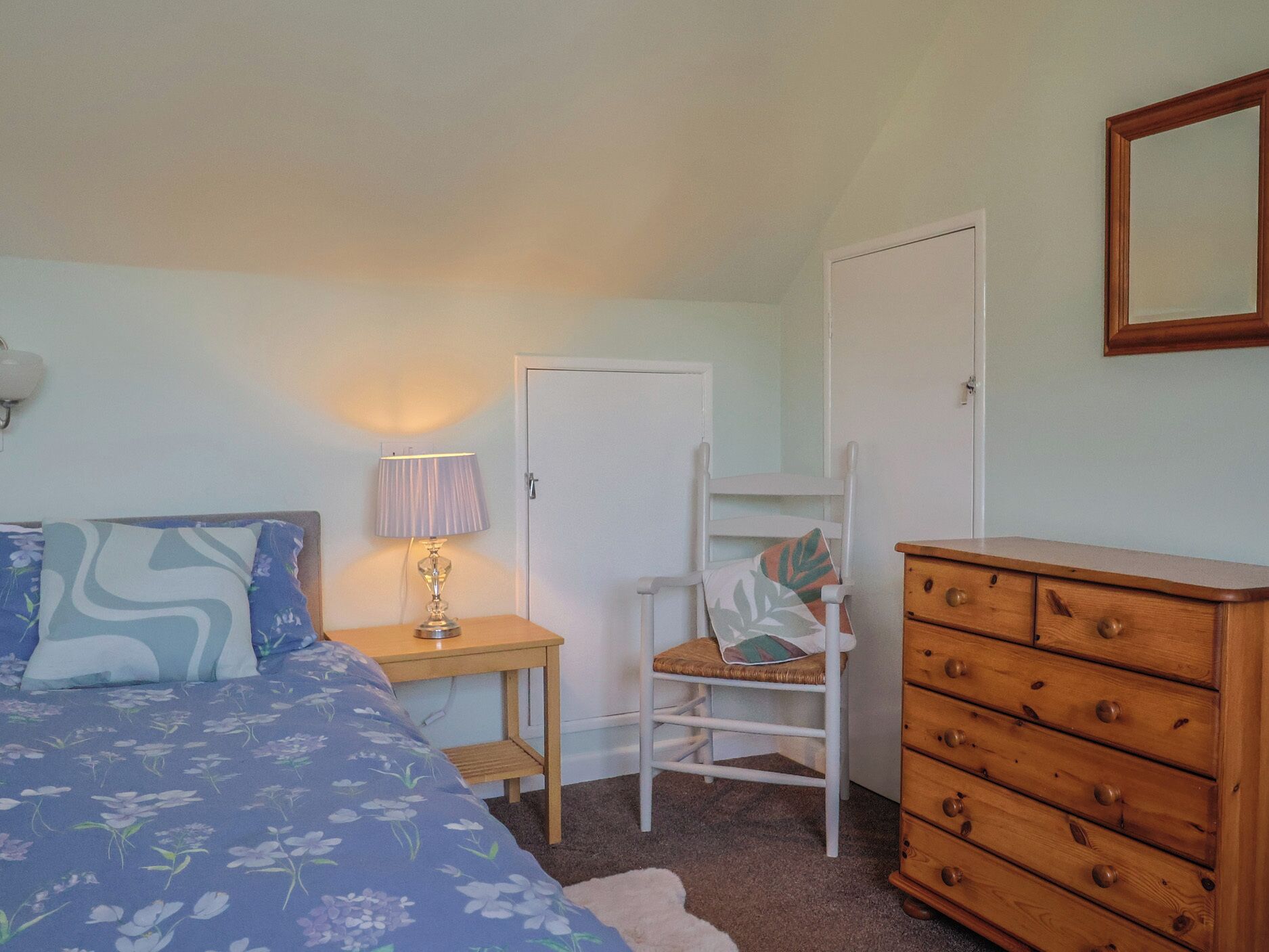 Un dormitorio con cama, una silla, cómoda, lámpara y una puerta.