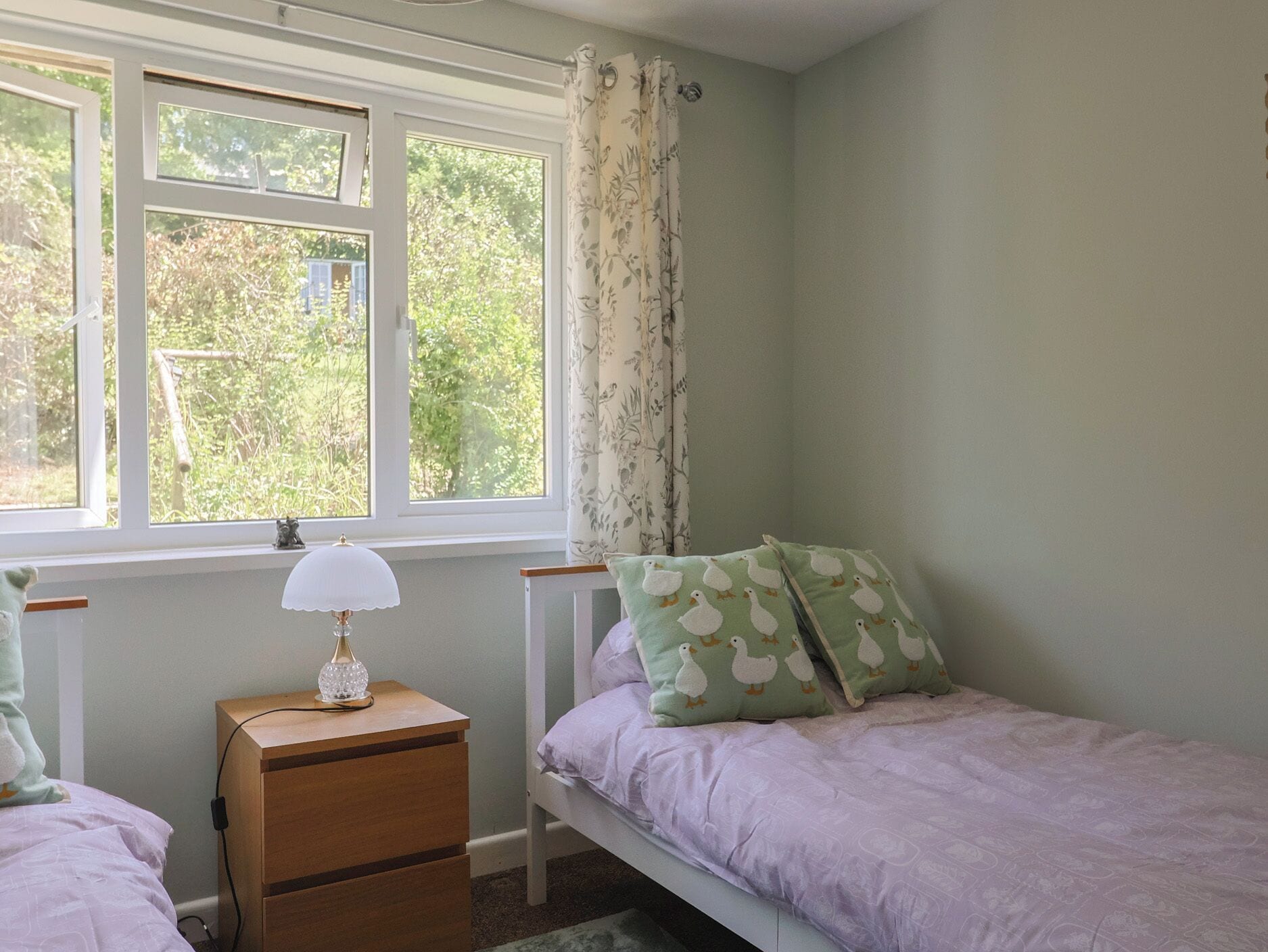 A bedroom with a single bed, a bedside table with a lamp, a window with curtains, and a view of greenery outside.