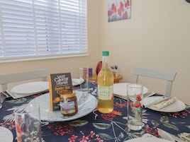 Una mesa de comedor con mantel floral, un libro titulado 'West Country Legends', una botella de jugo de manzana y un tarro de miel.