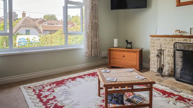A living room with a fireplace, a television mounted on the wall, a window with a view of houses, and a small table with magazines. - Tane (Blandford Forum)