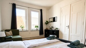 A hotel room with two beds, a green armchair, a desk, a mirror, and a window with curtains.