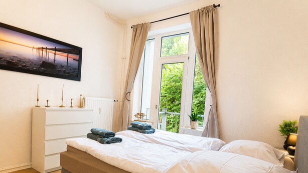 A bedroom with a bed, a dresser, a window with curtains, and a view of greenery outside.