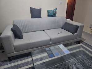 Un sofá gris con dos almohadas gris oscuro y una almohada floral de colores sobre una pared lisa. Un control remoto está sobre el sofá.