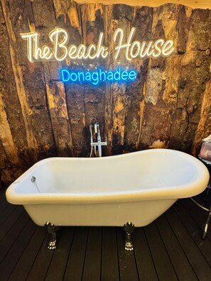 A white freestanding bathtub with a faucet, set against a wooden backdrop with a neon sign reading 'The Beach House Donaghadee.' - Posh 3-BDR Home, Beach House - Sleep6 - WiFi - Hot Tub - Free On-Street Parking (Donaghadee)
