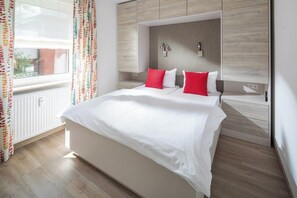 A modern bedroom with a large bed, two bedside tables, a headboard with built-in lighting, and a window with curtains. - // MeerZeit Norderney Vacation Rental // (Norderney)