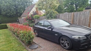 Um carro preto estacionado numa entrada de garagem com um caminho de tijolos, uma cerca de madeira e um jardim com flores vermelhas.