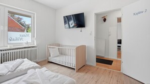Une chambre moderne et bien éclairée, équipée d’un berceau, d’un téléviseur fixé au mur et d’une fenêtre donnant sur un immeuble en briques.