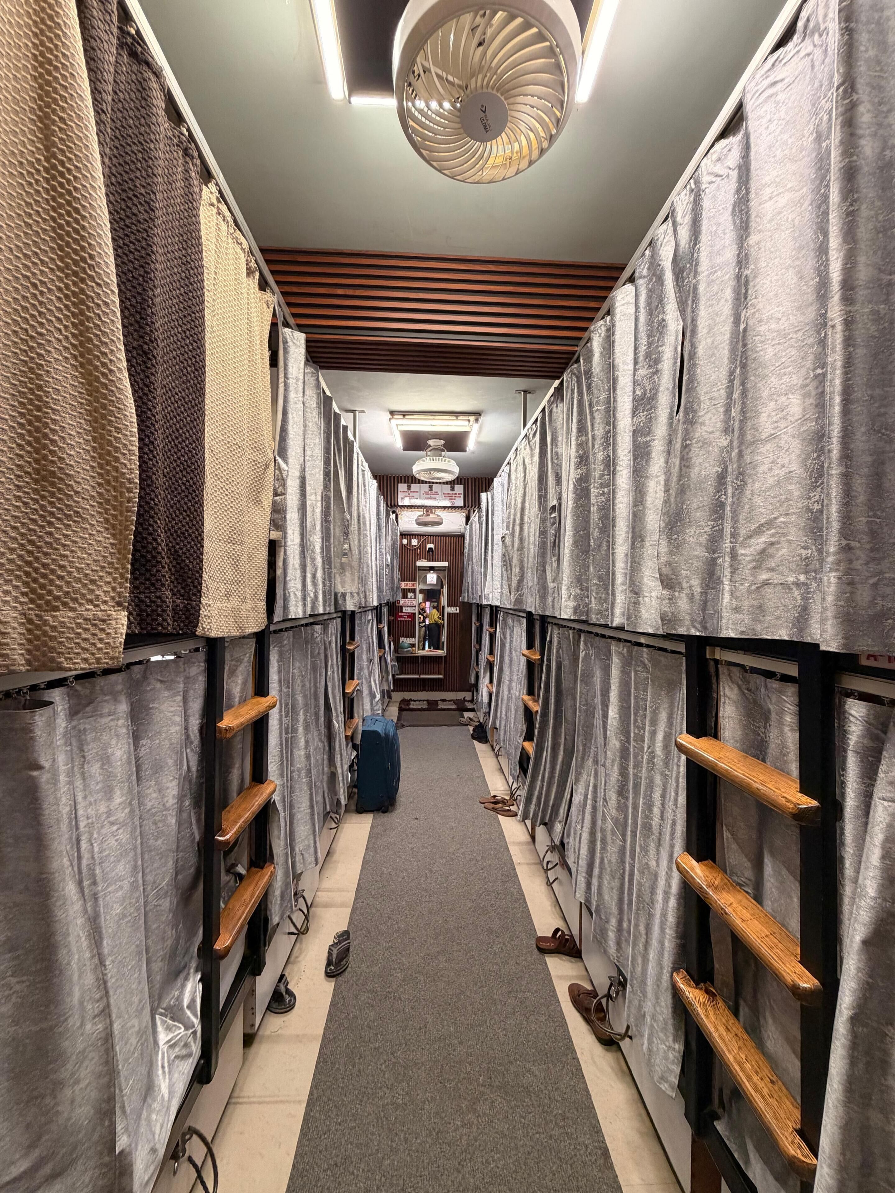 Un couloir étroit, bordé de rangées de lits recouverts de draps gris, avec des étagères en bois et un ventilateur au plafond.
