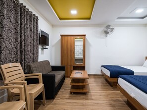A room with a sofa, a wooden table, a bed, a TV mounted on the wall, and a ceiling fan. - Vasu Inn - Rooms in Valparai (Valparai)