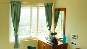 A room with a wooden desk, a laptop, a mirror, and a view of a green landscape through the window. - Vasu Inn - Rooms in Valparai (Valparai)