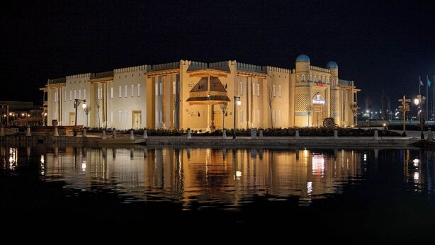 A large, illuminated building with a central entrance, surrounded by a reflective water body. - Arda Karvon Saroy (Khiva)