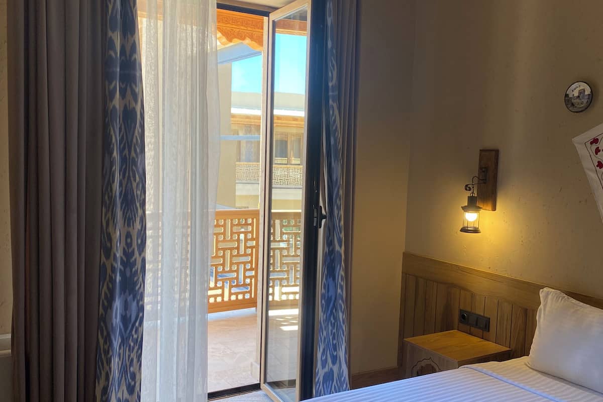 Quarto de hotel com cama, cortinas, mesa de cabeceira, luminária na parede e vista através de uma porta de vidro.