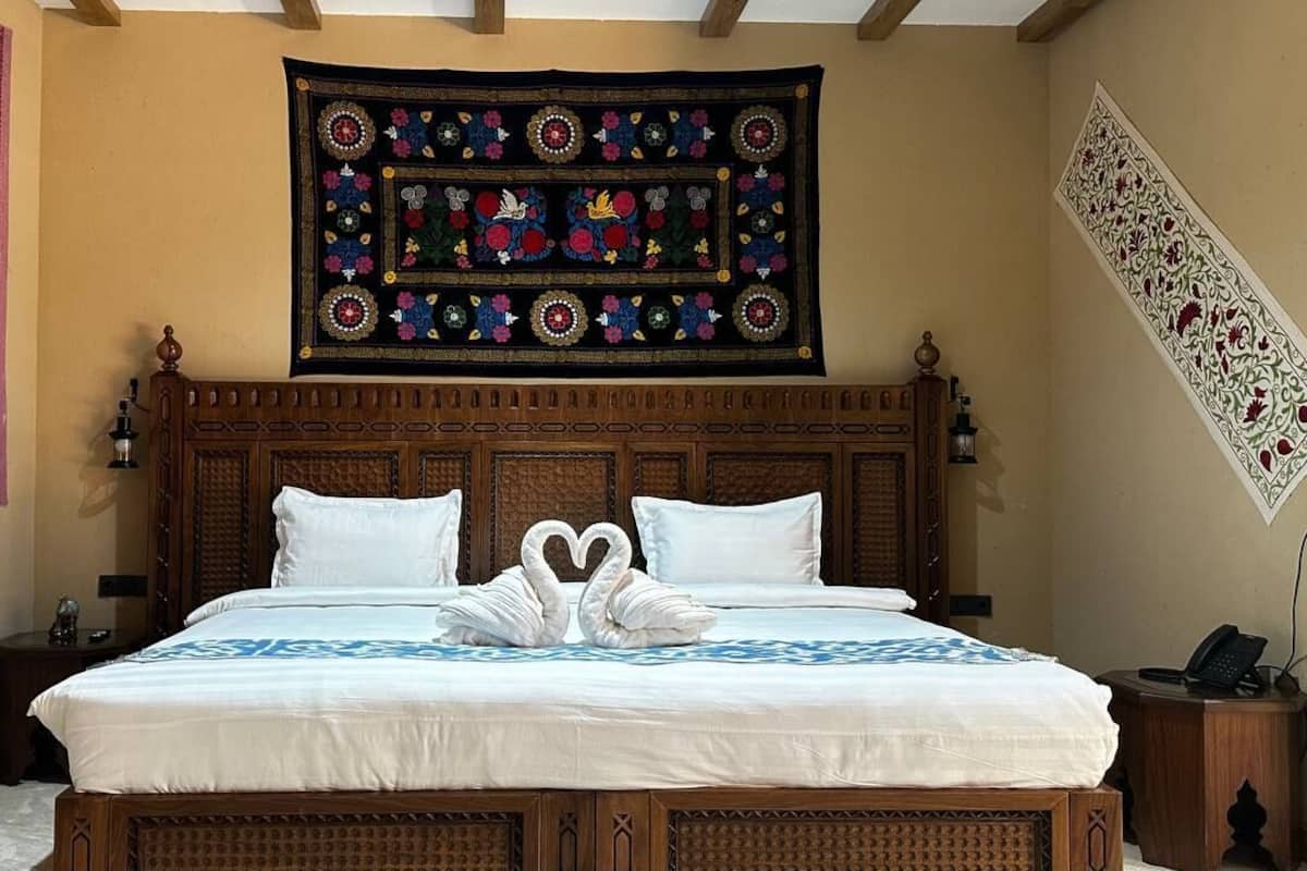 Uma cama com colcha branca e duas almofadas, cabeceira de madeira, tapeçaria acima da cama e mesa de cabeceira com um telefone.