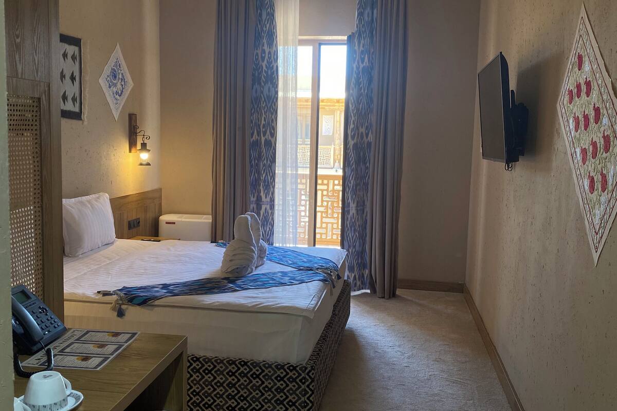 Quarto de hotel com cama, mesa de cabeceira, escrivaninha e televisão fixada na parede.