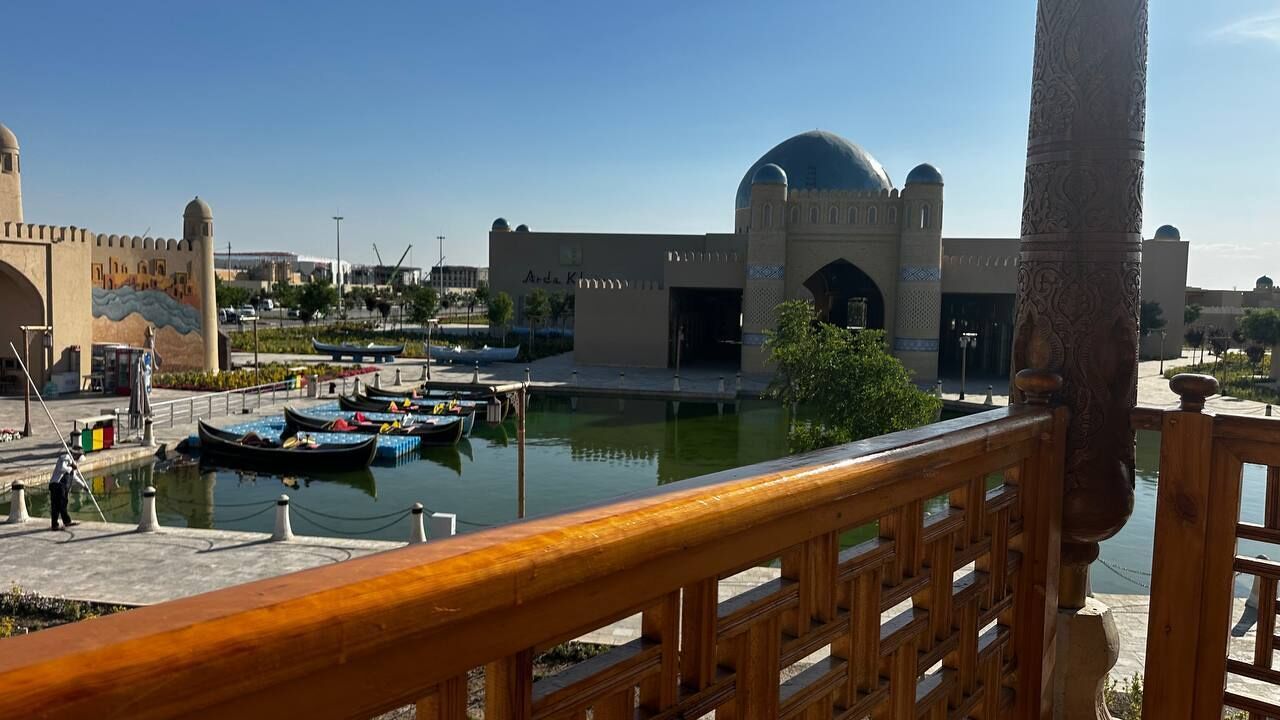 Una zona sul lungomare con un edificio che presenta una cupola e degli archi, una ringhiera in legno e barche ormeggiate.