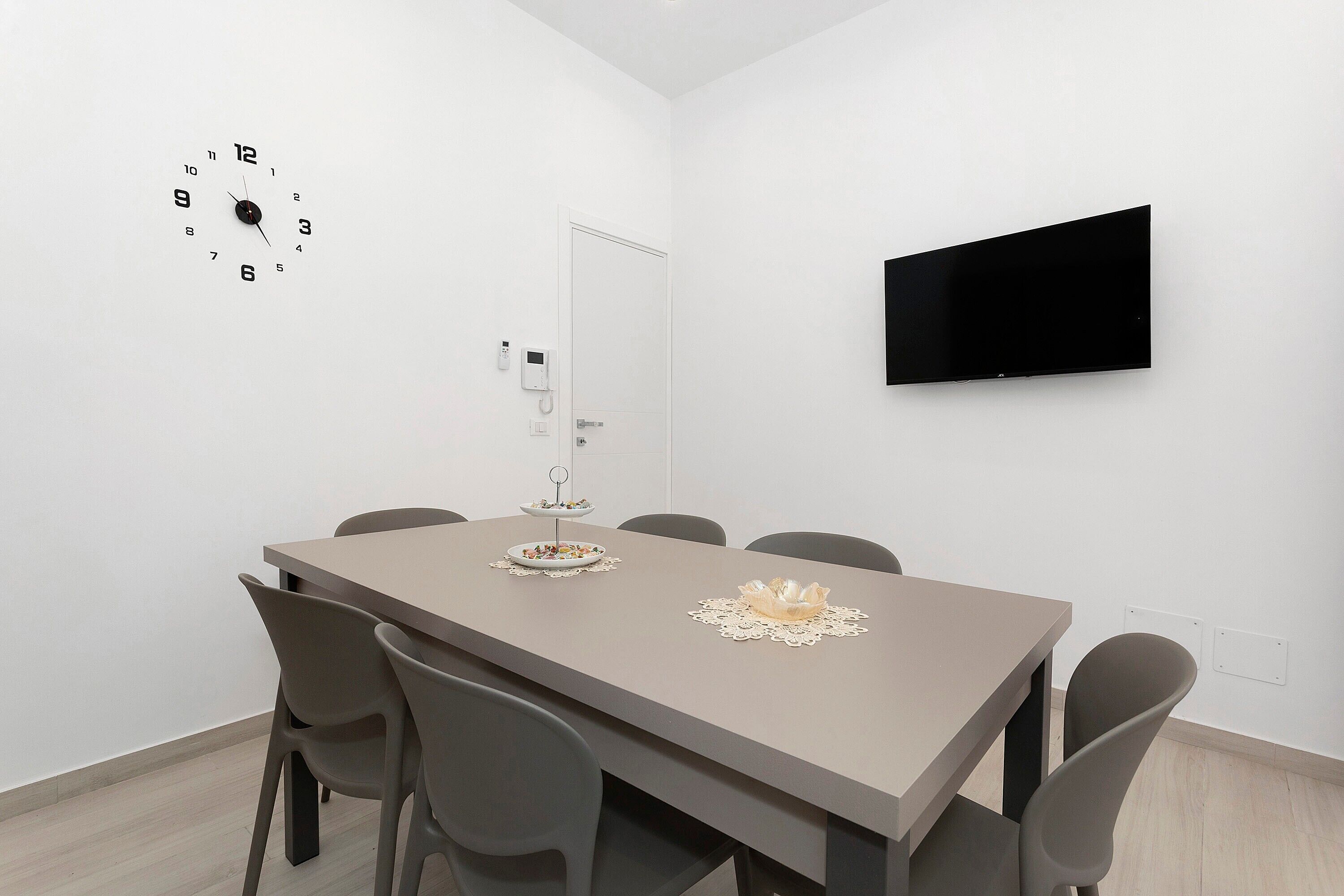 Uma sala de conferências com uma mesa, cadeiras, um relógio, uma porta e uma TV fixada na parede.