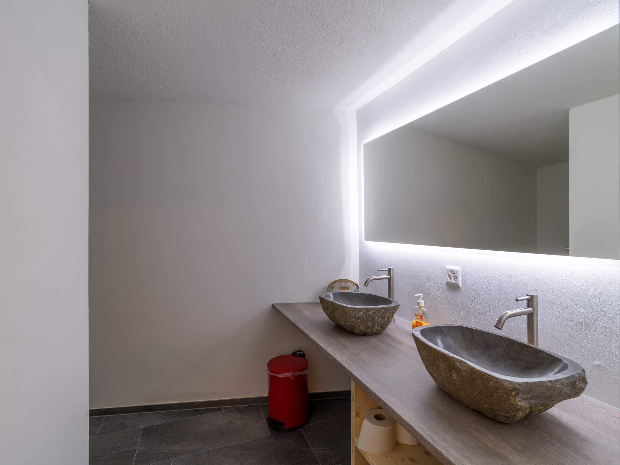 Un baño moderno con dos lavamanos de piedra, un cubo de basura rojo y un espejo grande.