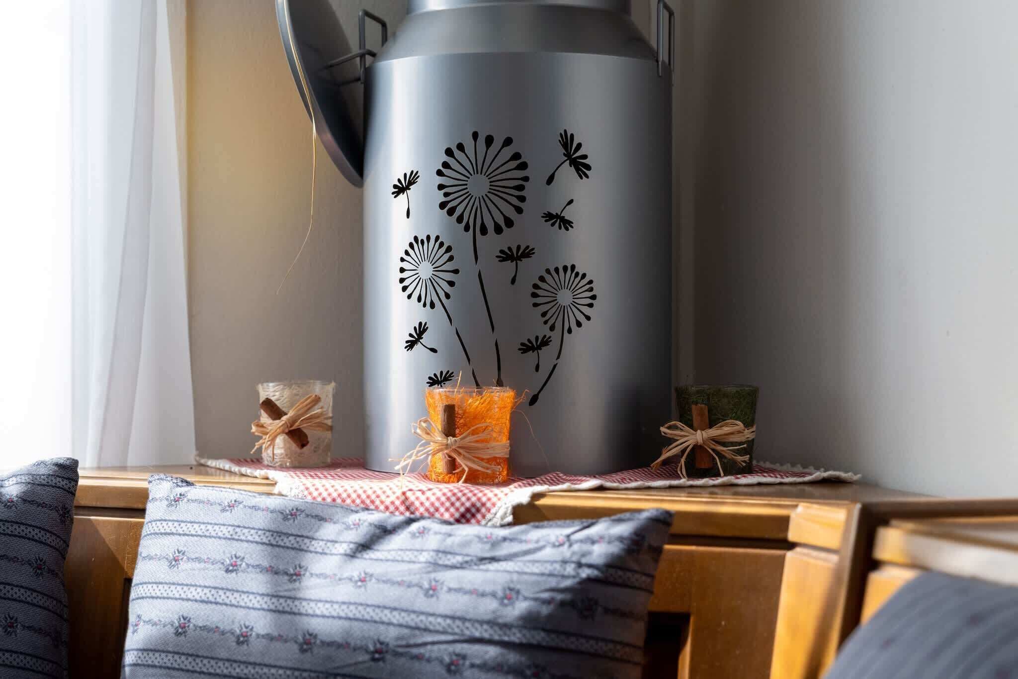 A metallic container with dandelion patterns, a glass jar with a cinnamon stick, and a checkered cloth on a wooden surface.