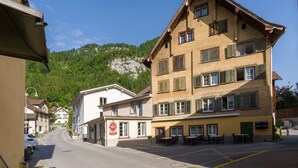 Une rue bordée d’immeubles, un restaurant avec une terrasse, et une montagne en arrière-plan.