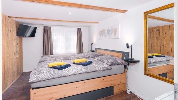 Une chambre à coucher avec un lit en bois, un téléviseur à écran plat, un miroir et une fenêtre avec des rideaux.
