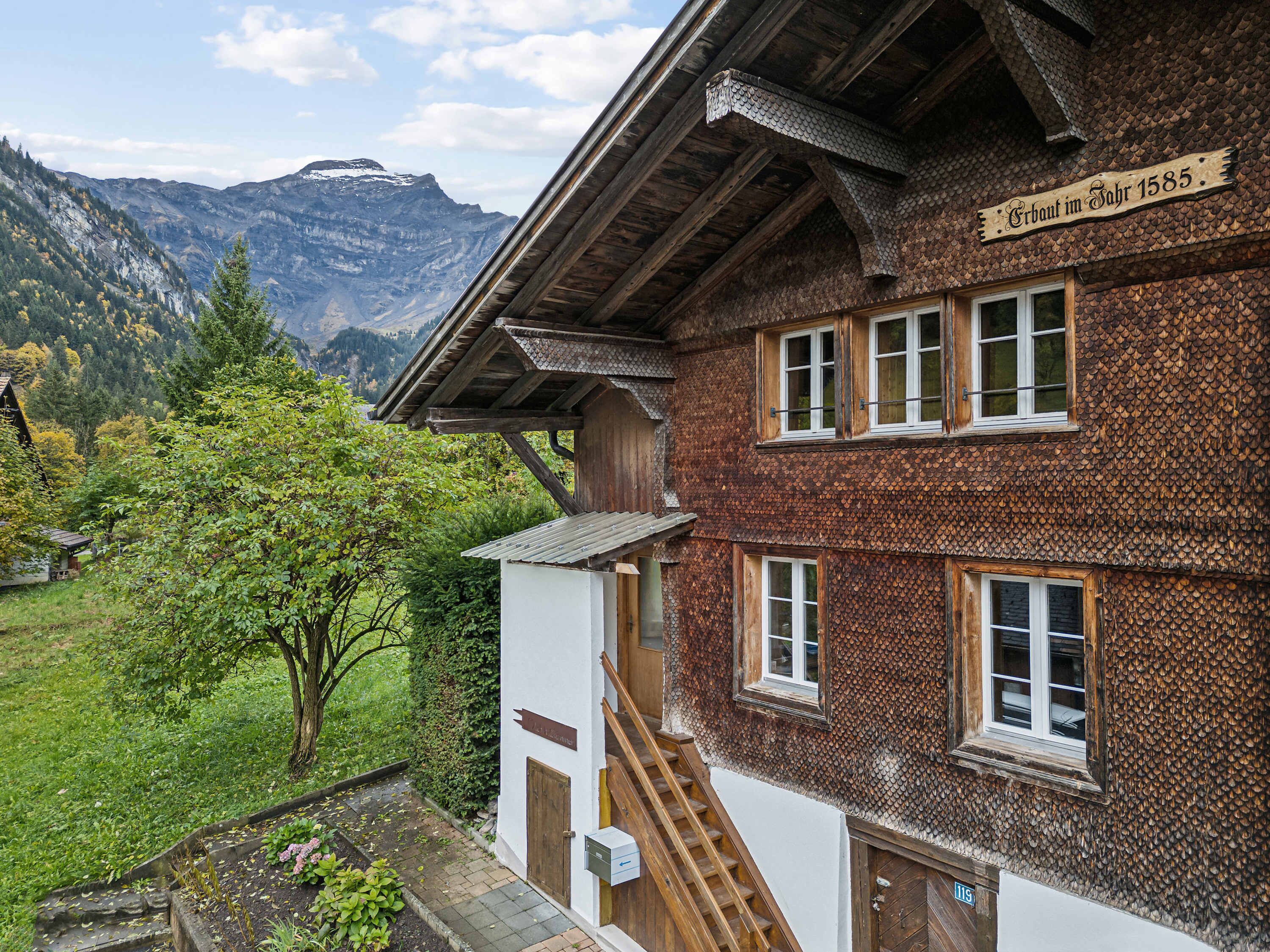 A traditional wooden building with a sign indicating its historical significance, a staircase leading to the entrance, and a view of a mountainous landscape.