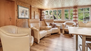 A room with a wooden ceiling, a sofa, a chair, a table, and large windows with curtains.