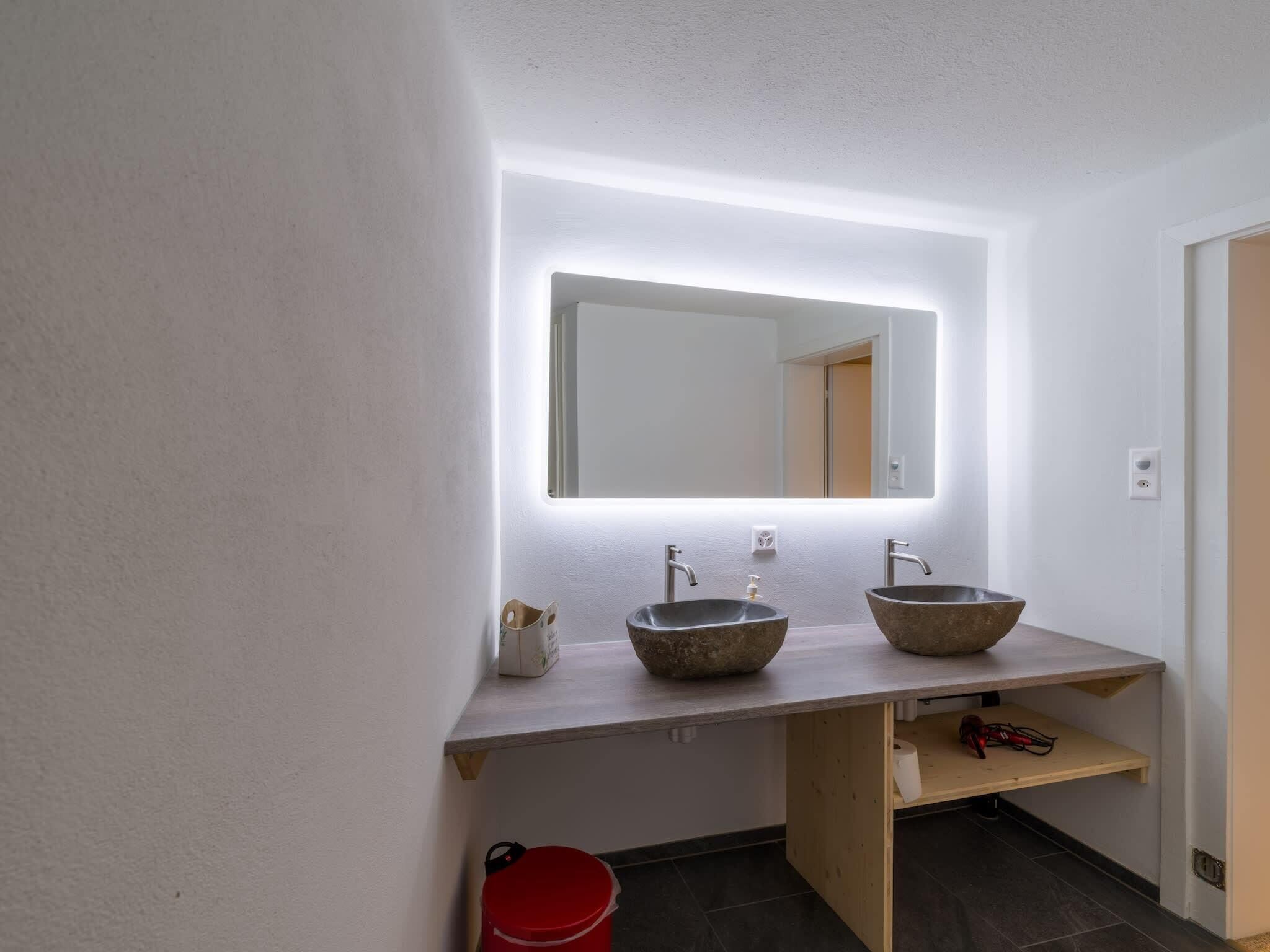 Un baño con un tocador blanco, dos lavamanos de piedra, un espejo con iluminación debajo del mueble y un cubo rojo.