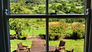 A window with a four-pane view of a green landscape, a wooden table with chairs, and a clear sky. - Scenic Retreat for 8 + Hot Tub with views over lakes, mountains & the sea. (Pentrefelin)