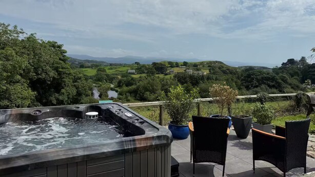 A hot tub on a patio with wicker chairs and potted plants, overlooking a green landscape with hills and a clear sky. - Scenic Retreat for 8 + Hot Tub with views over lakes, mountains & the sea. (Pentrefelin)