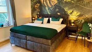 Un dormitorio con cama, una mesita de noche, una silla, una ventana con cortinas y un mural de hojas tropicales en la pared.