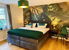 Ein Schlafzimmer mit einem Bett, einem Nachttisch, einem Sessel, einem Fenster mit Vorhängen sowie einer Wand mit einem tropischen Blatt-Wandgemälde.