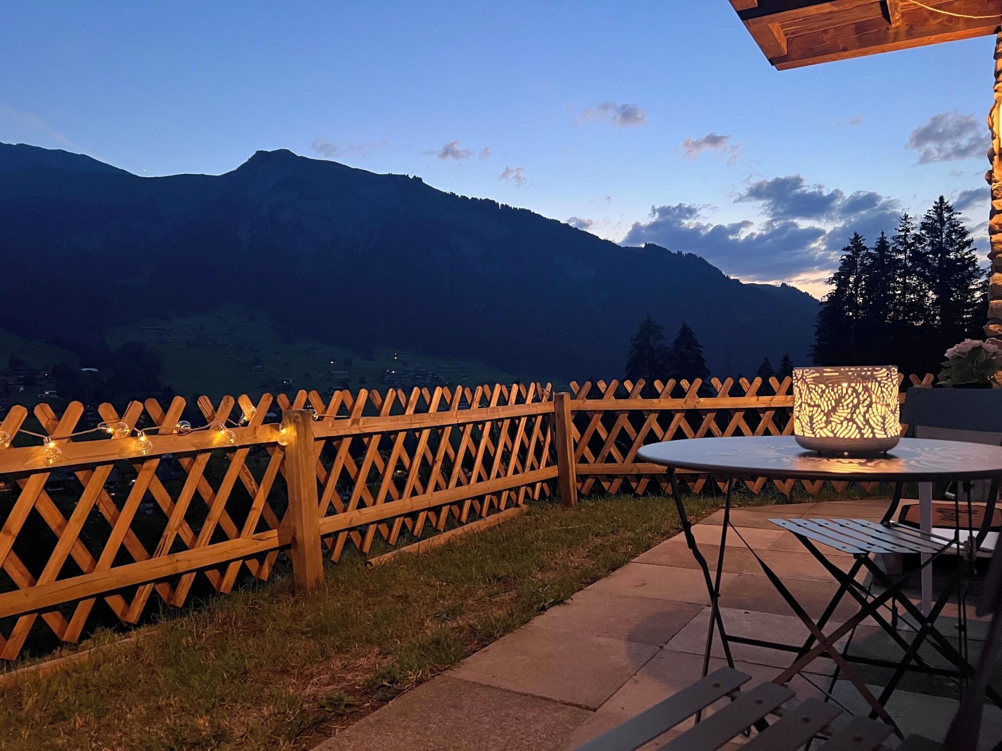 A patio with a wooden lattice fence, a table, and chairs, overlooking a mountain range.
