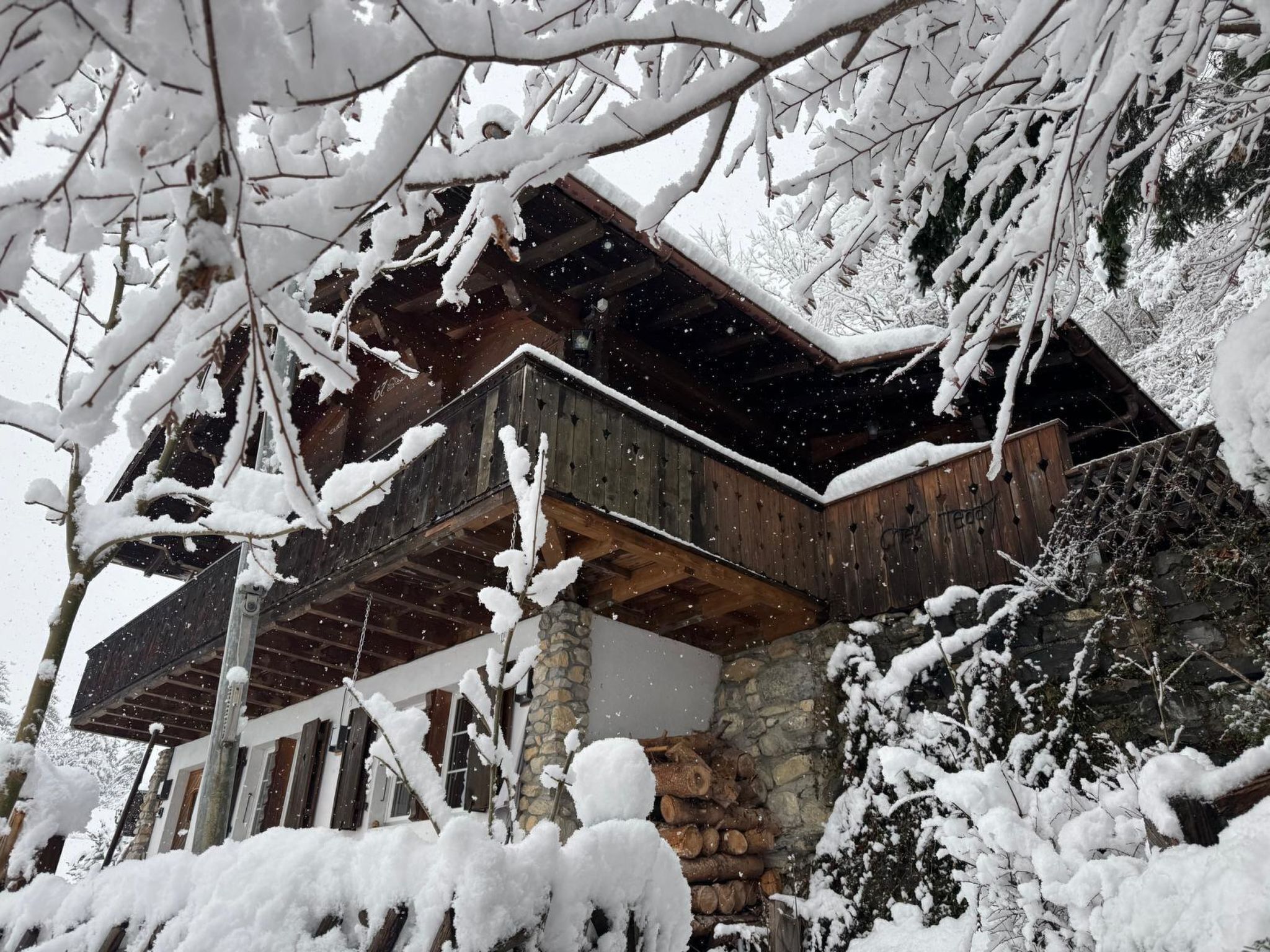 Una cabaña rústica con terraza de madera, paredes de piedra y ramas cubiertas de nieve.