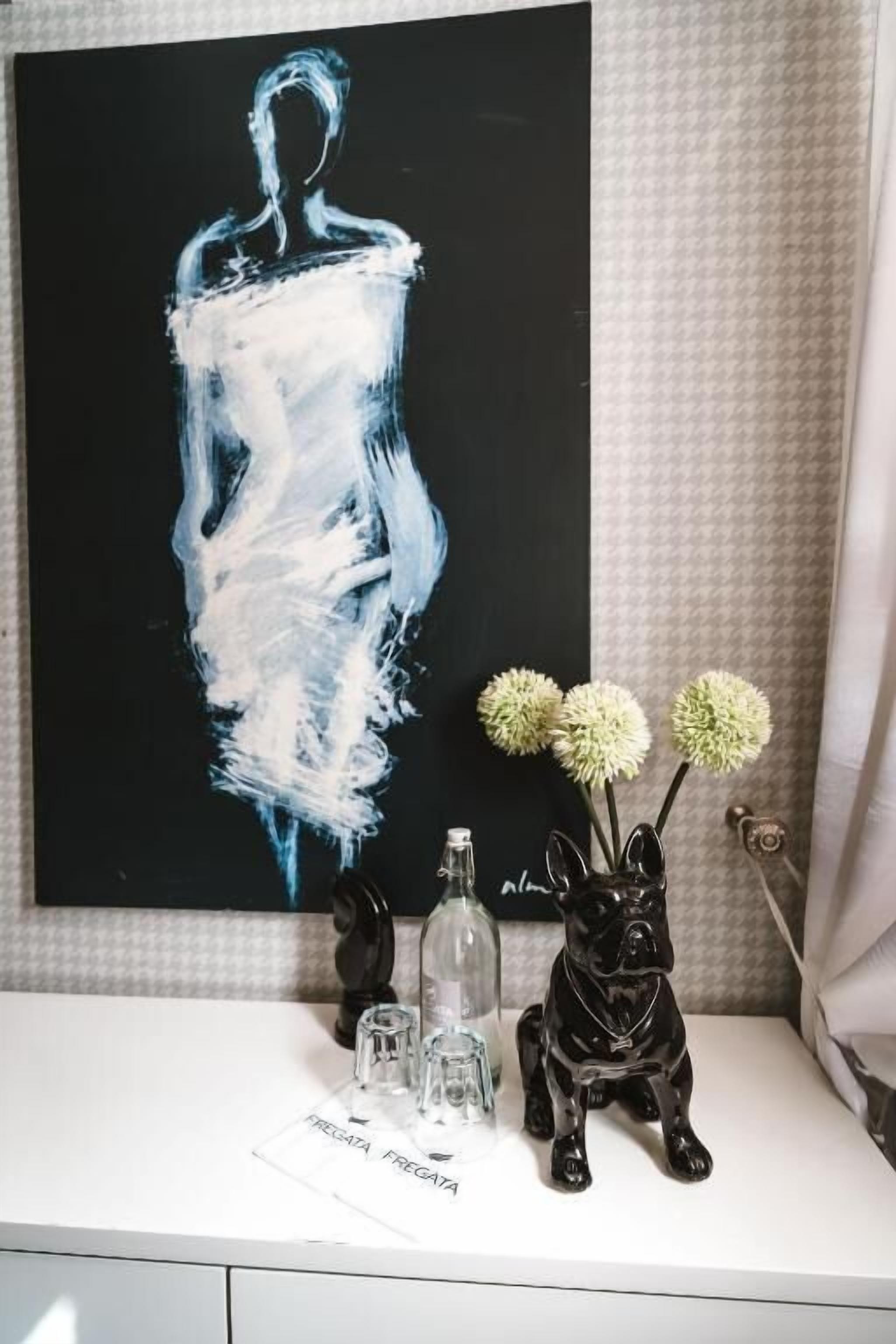 Une table blanche ornée d’un tableau en noir et blanc, un vase en verre contenant des fleurs, une bouteille en verre et une statuette noire.