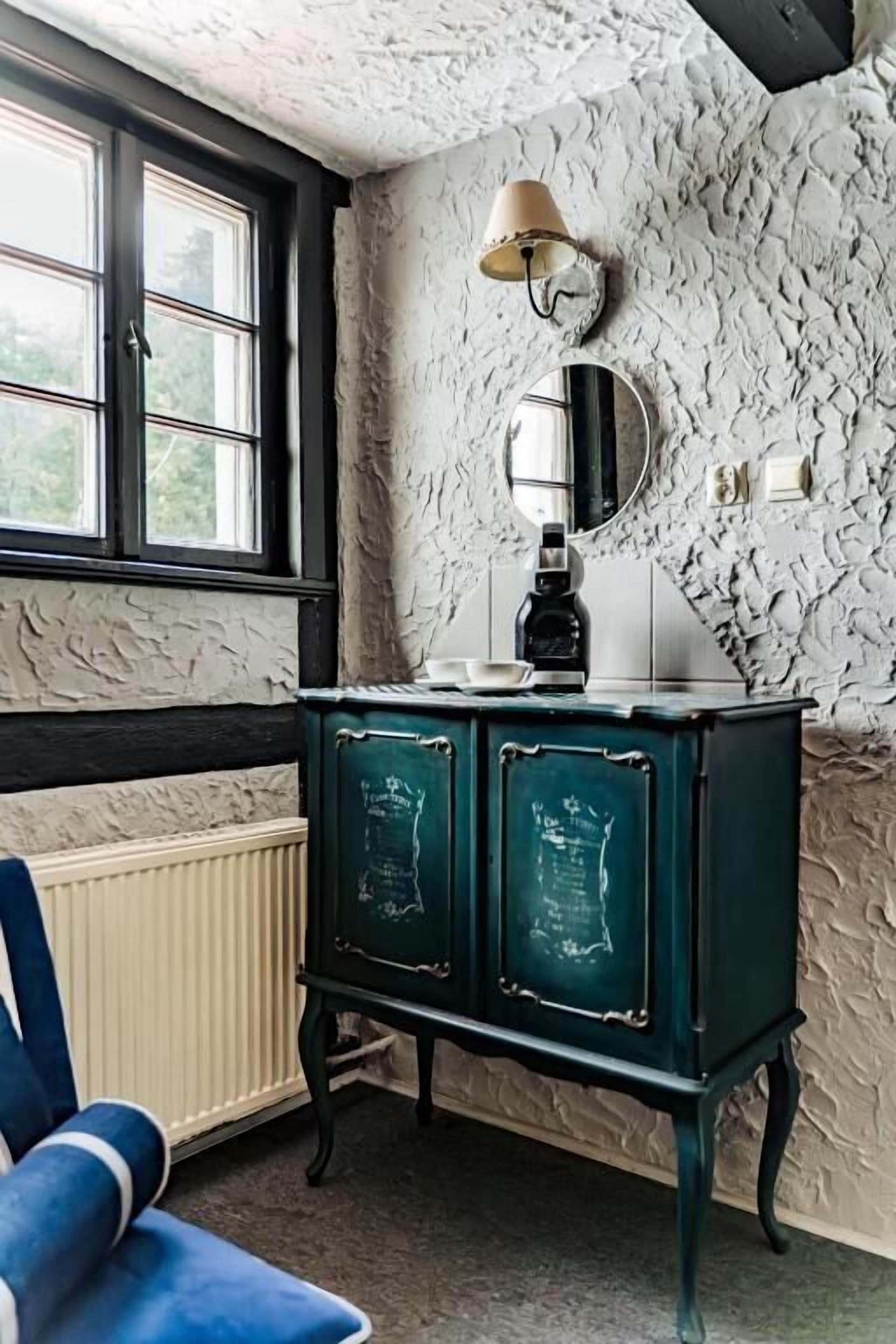 A room with a radiator, a blue chair, a green cabinet with chalkboard-style doors, a round mirror, and a window with a view of greenery.