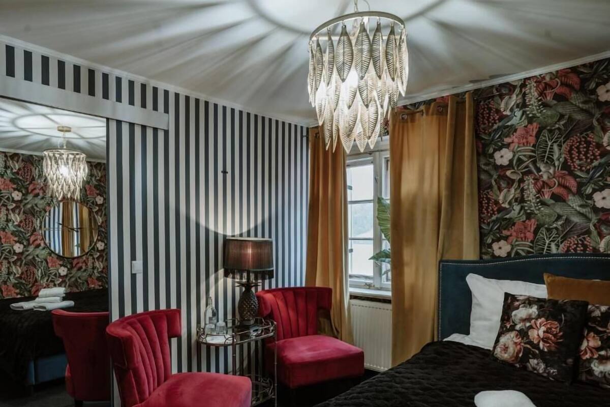 A hotel room with a bed, a red chair, a small table with a lamp, and a large window with curtains.