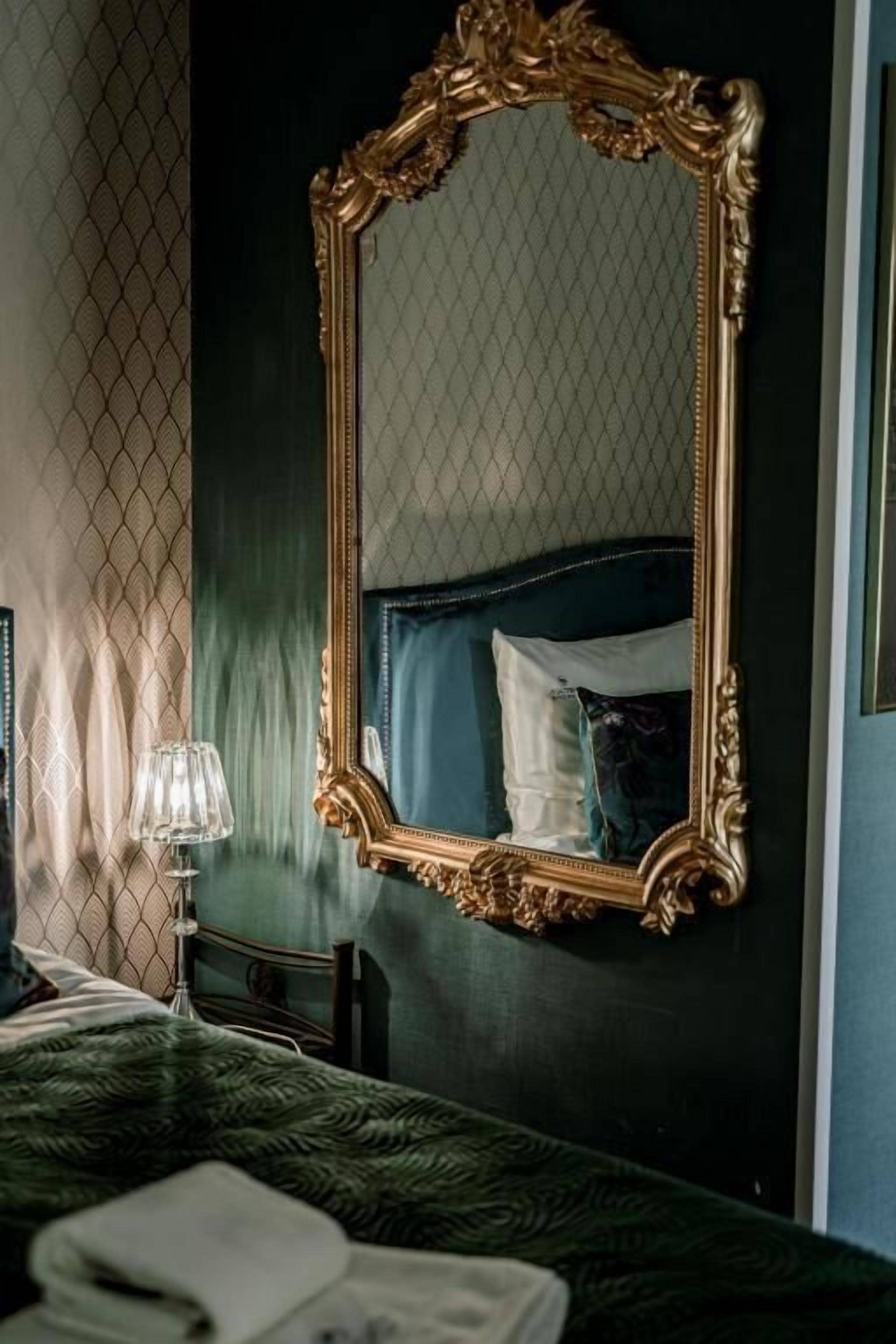 Una habitación con un espejo grande y ornamentado, una cama con colcha verde estampada, una mesita de noche con una lámpara y una ventana con cortina estampada.