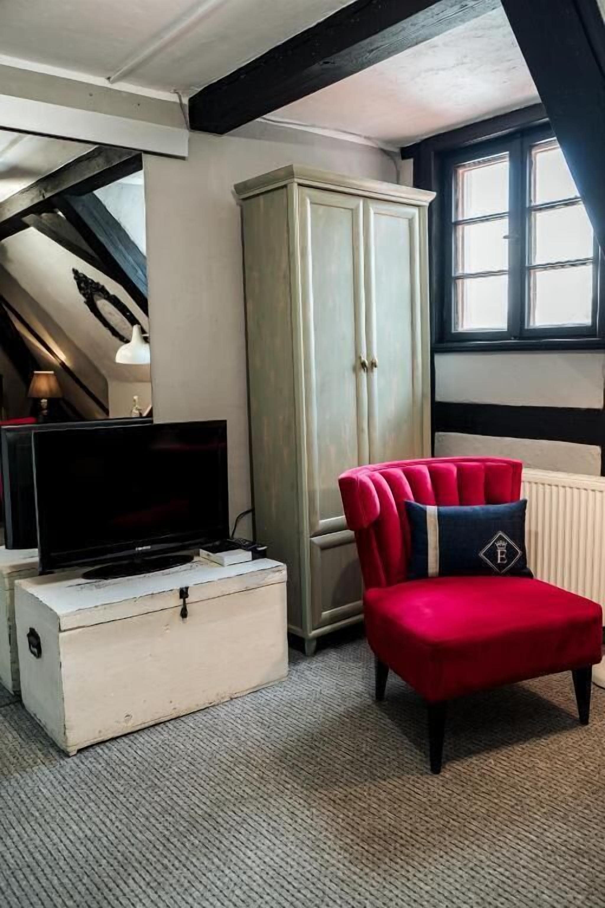 A room with a television on a dresser, a red chair with a pillow, a tall cabinet, and a window.