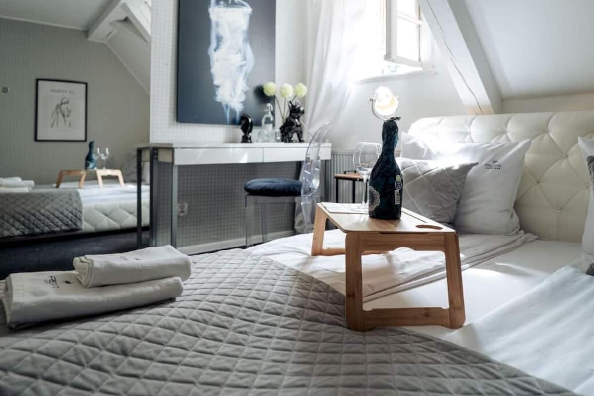A bedroom with a bed, a wooden nightstand, a desk, a chair, and a window.