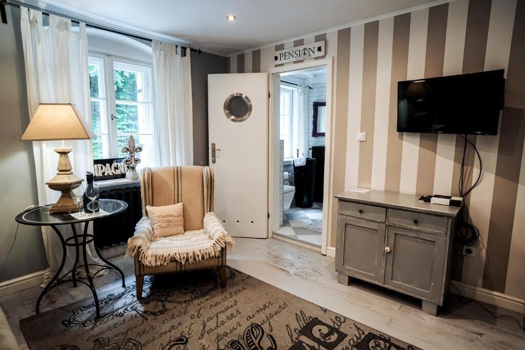 A room with a striped wall, a television mounted on the wall, a door labeled 'PENSION,' a small table with a lamp, and a chair.