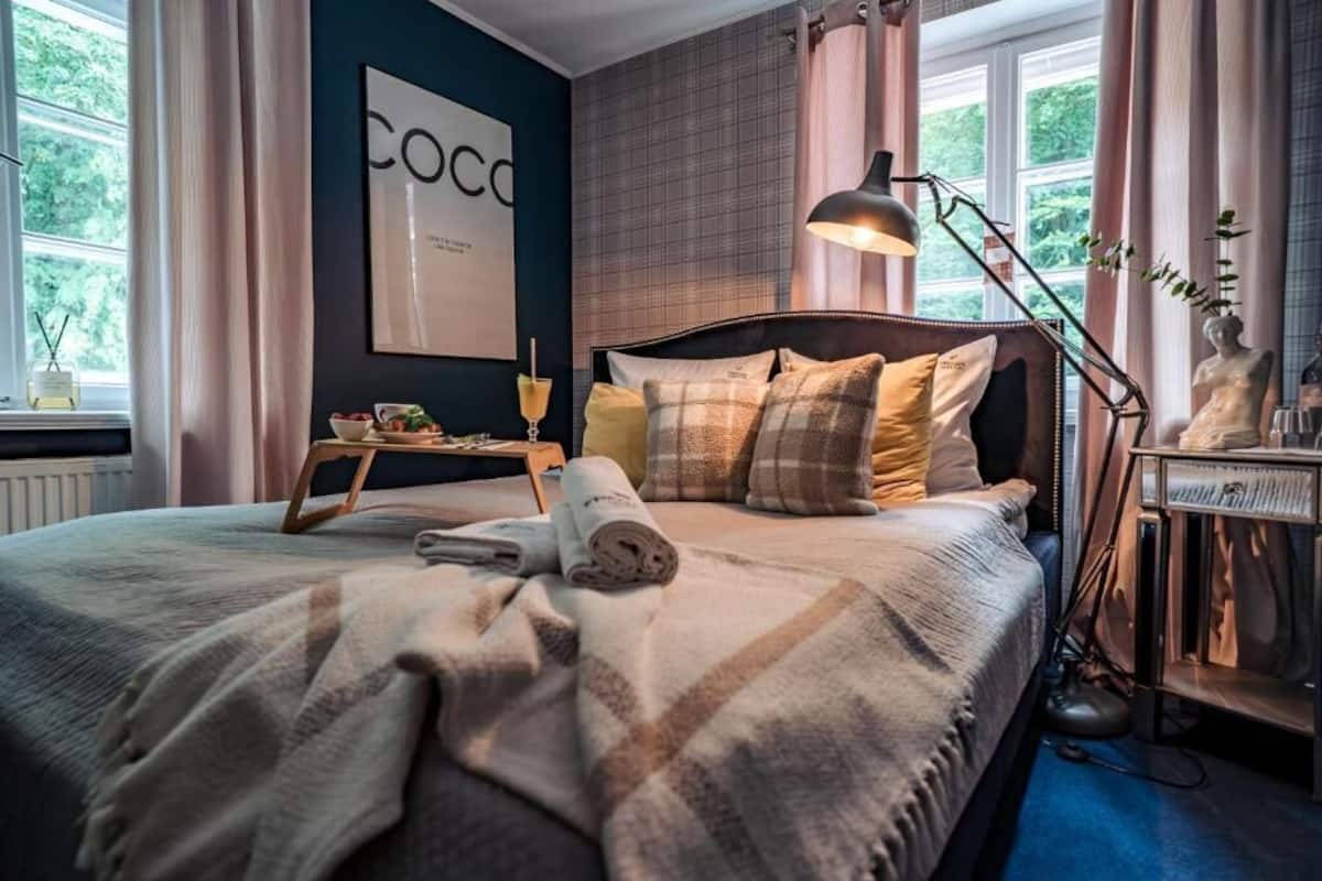 A bedroom with a bed, a bedside table, a lamp, a window, and a wall with a framed picture.