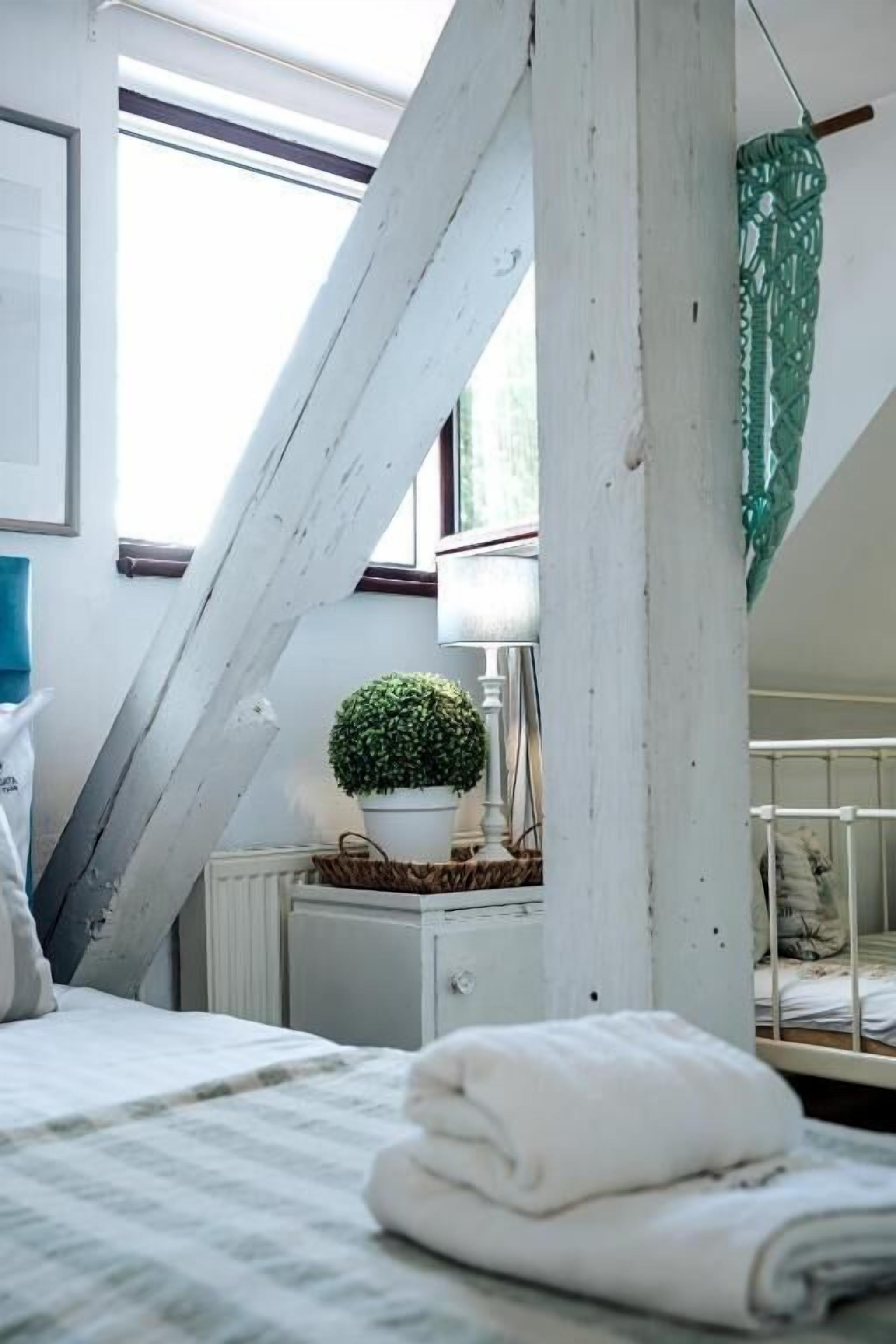 Un dormitorio con techo inclinado, una cama, una mesita de noche con lámpara, una pequeña planta en maceta y una ventana.
