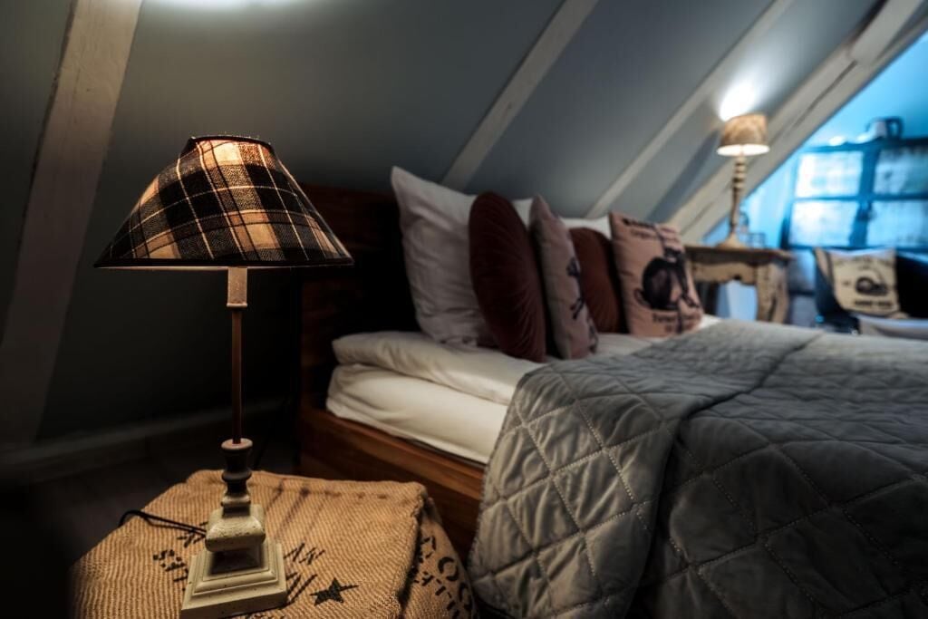 Ett sovrum med en säng, kuddar, ett vadderat täcke, en lampa och ett litet bord med en lampa på.